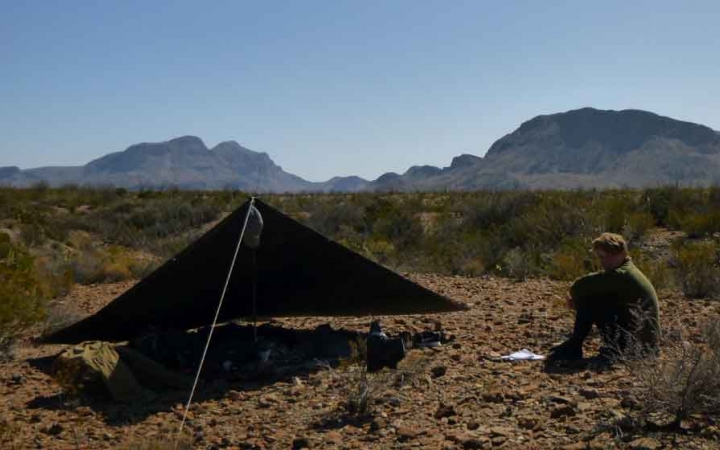 a student sits beside their tarp shelter in the desert with mountains in the background
